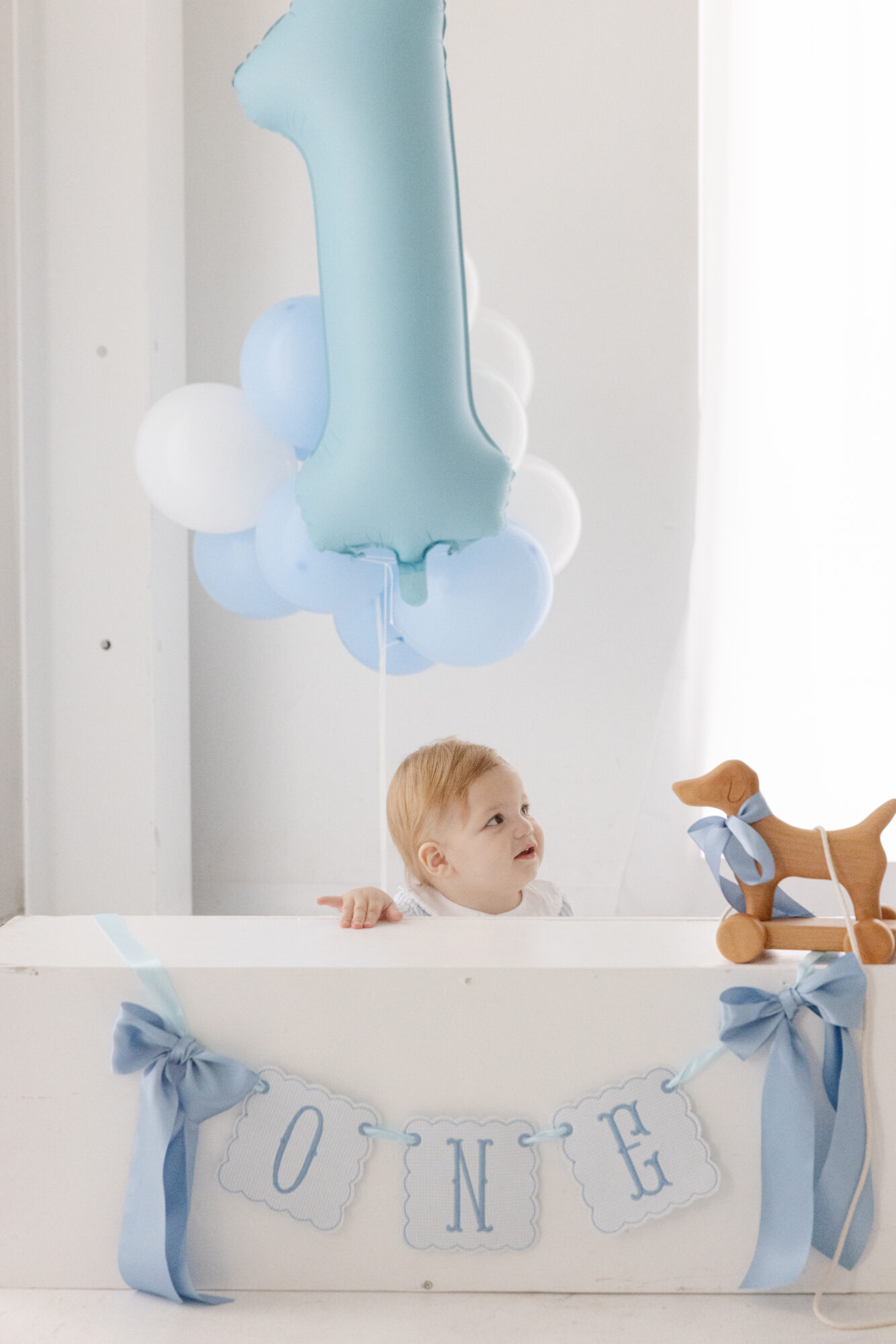 Birthday setup with blue balloons and a ONE banner as a baby boy peeks over the backdrop in a Houston studio.