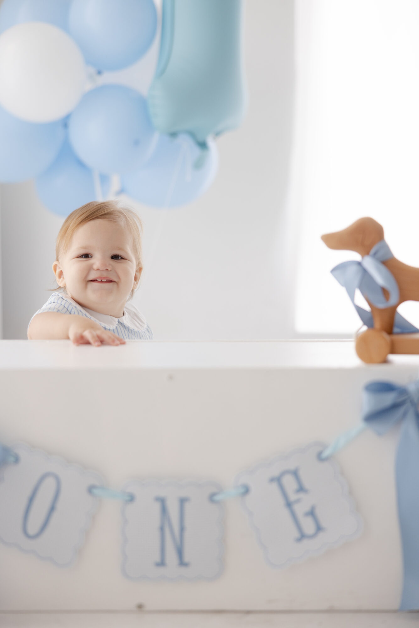 One year old boy smiling beside a blue balloons and ONE banner in a Houston photography studio.